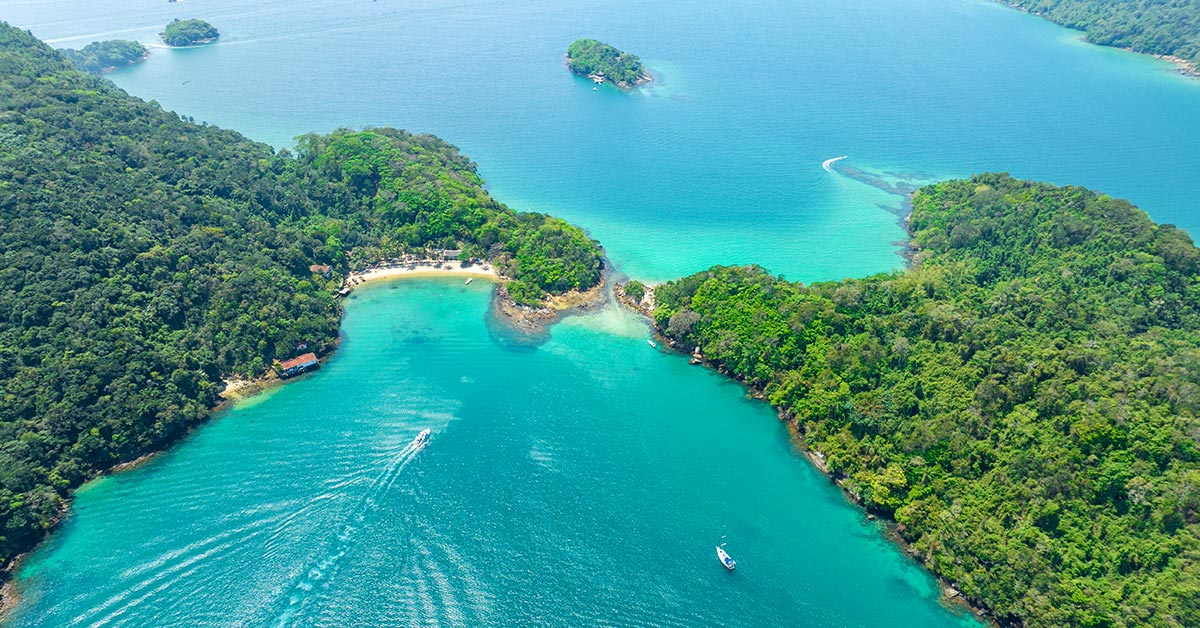 Vue aérienne d'une eau turquoise entre des îlots boisés, avec une petite plage et des bateaux près d'Ilha Grande